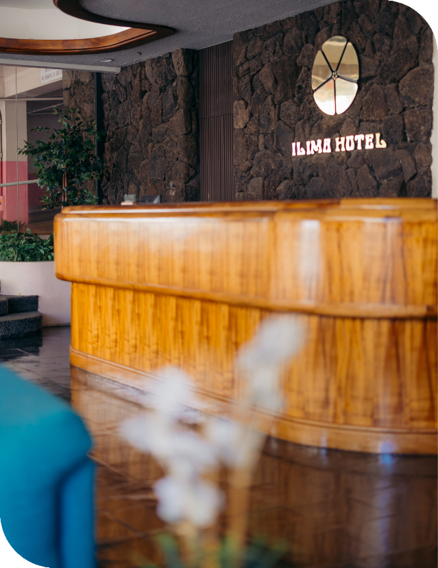 The Ilima Hotel lobby showcases a koa wood front desk illuminated by soft lighting, set against a striking lava rock wall